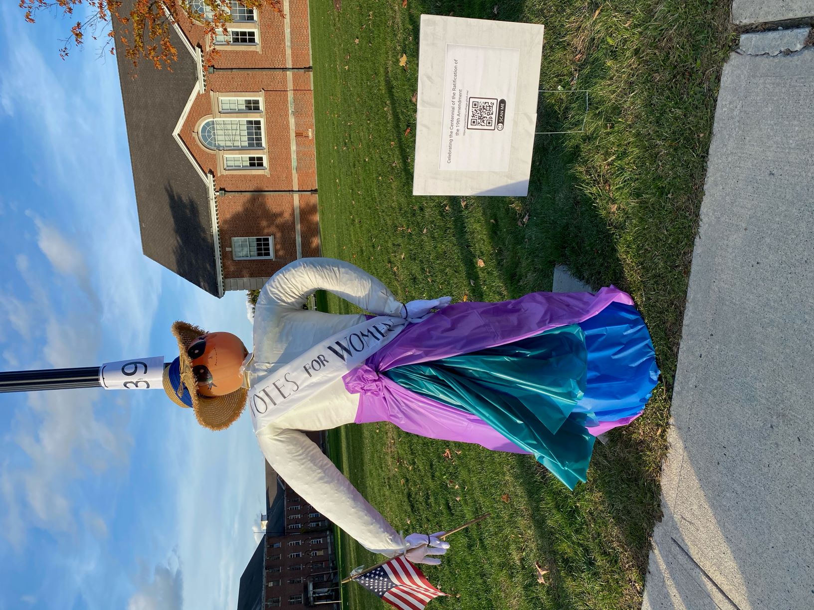 suffragette scarecrow wearing a big hat, white button down shirt, long skirt, and sash that says 'Votes for Women'